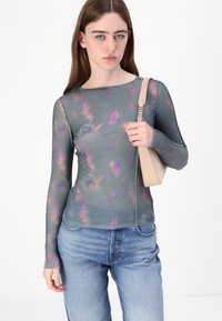 Long-sleeve, sheer top in grey with a floral pattern in purple and pink hues; paired with light blue, loosely fitting jeans and a beige bag.
