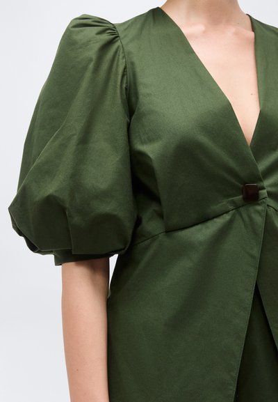 Γυναίκα που φοράει ένα ελιάς πράσινο φόρεμα με βαθύ V λαιμόκοψη, κοντά φουσκωτά μανίκια και ένα μαύρο κουμπί στη μέση.