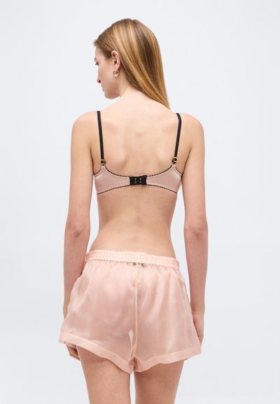 Γυναίκα με ίσια ξανθά μαλλιά φοράει ανοιχτό ροζ σουτιέν με μαύρα λουριά και διάφανα ροζ σορτς, στέκεται μπροστά σε ένα απλό ανοιχτό φόντο.
