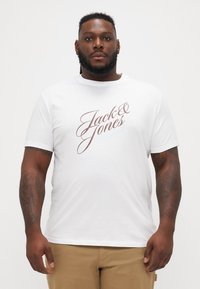 Bijela pamučna majica s kratkim rukavima koja sadrži ispisani logo "Jack & Jones" u smešnoj i plavoj boji. Okrugli izrez, standardna kroj.