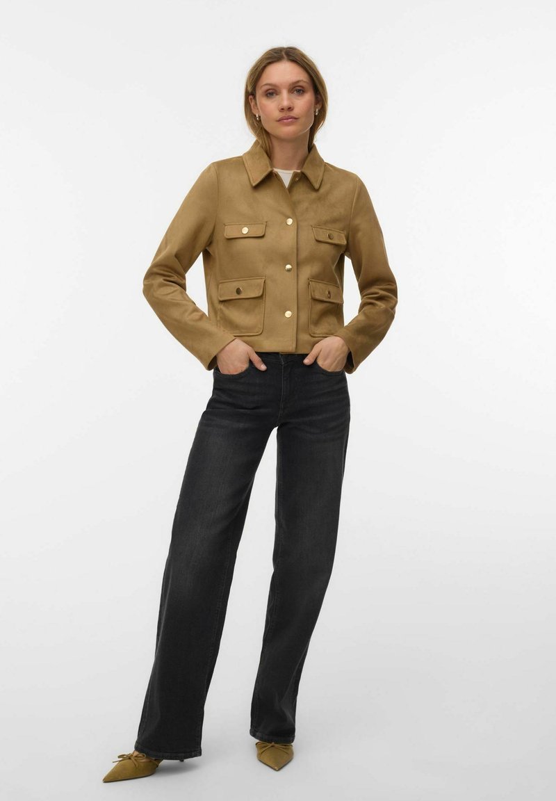 Femme portant une veste marron clair boutonnée avec quatre poches à l'avant, un jean noir taille haute à jambes droites, et des ballerines pointues marron clair, debout les mains dans les poches.