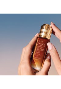 Bottiglia di vetro marrone con tappo dorato, etichettata "Estée Lauder Advanced Night Repair", tenuta in mano su uno sfondo sfocato.