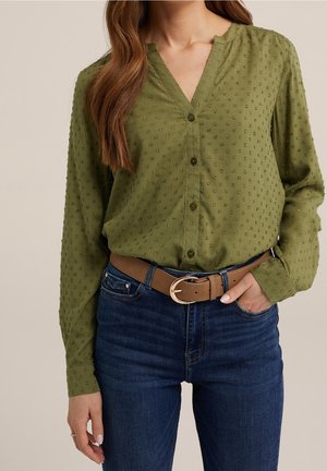 Olivengrøn bluse med et subtilt teksturmønster, v-hals og knaplukning, kombineret med højttaljede mørkeblå jeans og et tanfarvet bælte.