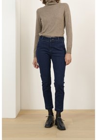 Pull à col roulé en maille beige, jean bleu foncé longueur cheville, chaussures noires à lacets et chaussettes noires à motifs sur fond neutre.