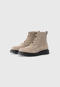 Stivaletti alla caviglia in suede beige con design a lacci, punta rotonda e suola in gomma nera. Presenta un logo discreto sul lato e una costruzione robusta.