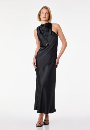 Femme aux cheveux blonds portant une robe noire sans manches en satin et des sandales noires à talons hauts, debout sur un fond blanc.