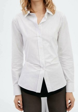 Kobieta ubrana w dopasowaną białą koszulę w delikatne prążki zapinaną na guziki z długimi rękawami oraz czarne szorty pod spodem.