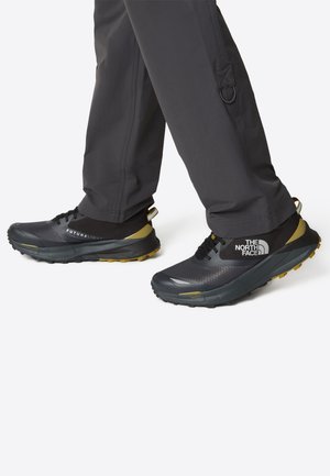 Scarpe da trail running nere e grigie con tomaia testurizzata, suola gialla e logo a contrasto. Design leggero con lacci e colli imbottiti.