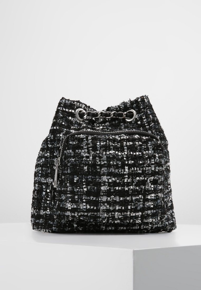 Bolsa de tela tejida en negro y plata con cierre de cordón, que cuenta con un bolsillo con cremallera en el frente y un acento de cadena en la parte superior.
