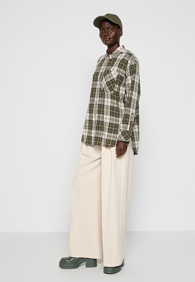 Camisa de cuadros verde con dos bolsillos en el pecho, corte oversized. Combinada con pantalones blancos holgados y zapatos gruesos de color verde oscuro. Incluye una gorra verde.