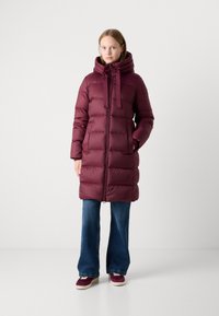 Bordeaux puffer coat met een capuchon, frontale rits, verstelbare trekkoorden en ribgebreide manchetten, gedragen over flared denim jeans en sneakers.