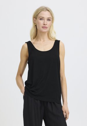 Femme blonde portant un haut noir sans manches et un pantalon noir, debout avec une main dans la poche, sur un fond clair uni.