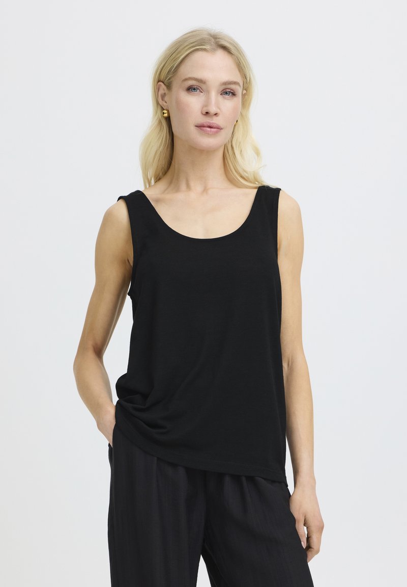 Femme blonde portant un haut noir sans manches et un pantalon noir, debout avec une main dans la poche, sur un fond clair uni.