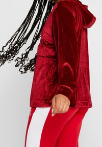 Sweat à capuche en velours rouge avec une finition texturée, doté d'une poche avant et d'une capuche. Porté avec un pantalon rouge assorti avec des bandes blanches sur les côtés.