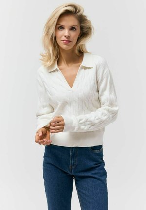 Blondynka z rozpuszczonymi włosami w białym dzianinowym swetrze i niebieskich dżinsach poprawia rękaw na jasnym, jednolitym tle.