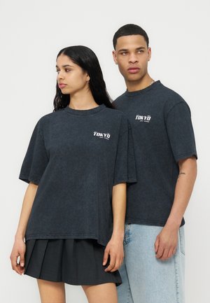 Δύο μοντέλα που φορούν σκούρα, φαρδιά T-shirts με σχέδιο Tokyo, το ένα συνδυασμένο με μαύρη φούστα με πιέτες και το άλλο με ανοιχτόχρωμο μπλε τζιν, στέκονται μπροστά σε λευκό φόντο.