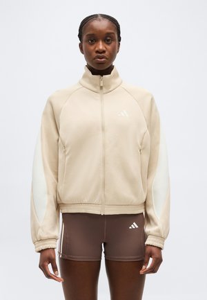 Junge Person trägt eine beige Jacke mit Reißverschluss, weißen Ärmelpanelen und braune Sportshorts mit Logos, steht vor einem schlichten Hintergrund.