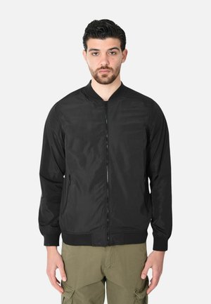 Uomo con capelli scuri e barba, indossa una giacca bomber nera con cerniera e pantaloni cargo verde oliva, in piedi contro uno sfondo chiaro uniforme.