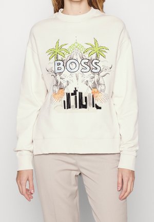 Biała bluza z nadrukiem „BOSS”, zielonymi palmami, kwiatami i stylizowaną grafiką miejskiego krajobrazu noszona przez stojącą osobę.