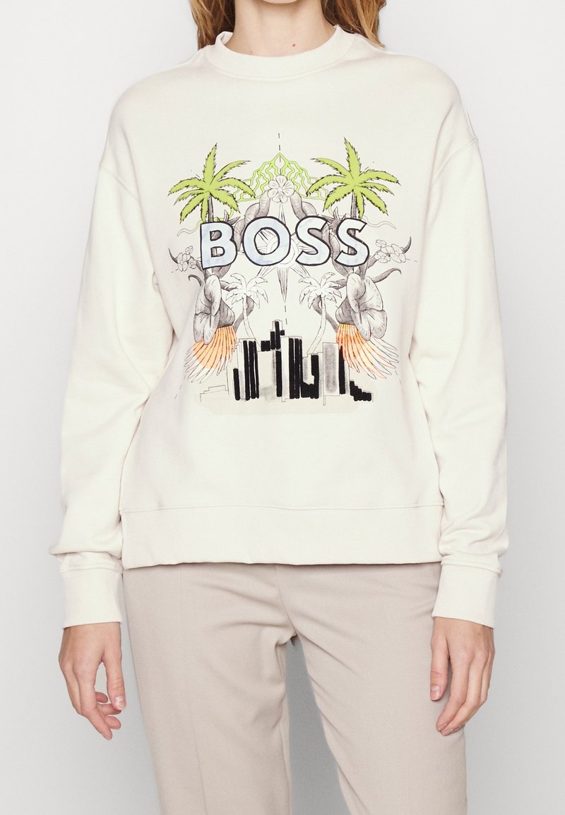 Sweat-shirt blanc avec le texte « BOSS », palmiers verts, fleurs et un graphisme stylisé d'un paysage urbain porté par une personne debout.