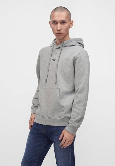 Homme portant un sweat à capuche gris clair avec poche avant et cordons, associé à un jean bleu foncé, debout devant un fond blanc uni.