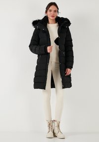 Abrigo de plumas negro con capucha de piel sintética, cierre delantero y bolsillos laterales. Lleva un suéter crema y pantalones beige claro con botas a juego.