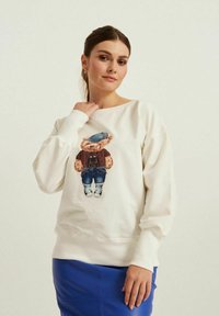 Biały sweter z grafiką niedźwiedzia w swobodnym stylu. Niedźwiedź ma na sobie brązową kurtkę, niebieskie dżinsy i sneakersy. Długie rękawy, luźny krój.