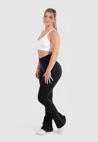 Zwarte uitlopende leggings en een wit crop topje. De stof lijkt rekbaar te zijn met een gladde textuur. Model poseert zijwaarts tegen een neutrale achtergrond.