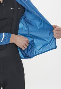 Blå vindjacka med dragkedja fram, elastiska ärmslut och meshinredning, med en sidoficka och en mjuk, lätt textur.