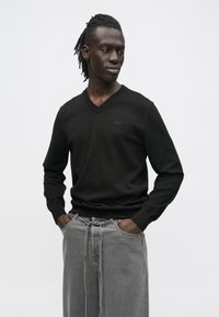 Fonott hajú férfi fekete V-nyakú pulóvert és laza szürke farmert visel, kezét a zsebében tartva, oldalra nézve, fehér háttér előtt.