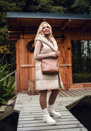 Gesteppte beige Weste, braunes Oberteil, braune Leggings und weiße Stiefeletten. Braune Schultertasche. Holzhütte und Steg in natürlicher Umgebung.