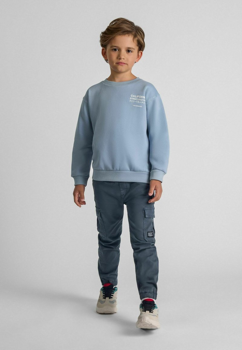 Ljusblå sweatshirt med ribbade muddar, prydd med vit text. Kombinerad med mörkgrå cargobyxor med elastiska muddar och flerfärgade sneakers.