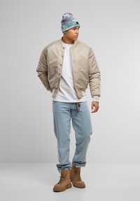 Giubbotto bomber beige con finitura liscia, t-shirt bianca, jeans azzurri chiari, stivali color sabbia e berretto di maglia tie-dye in blu e viola.