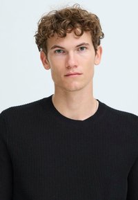 Schwarzer, gerippter Strickpullover mit rundem Halsausschnitt und strukturierter Oberfläche. Das Model hat helles, lockiges Haar und blauäugig.