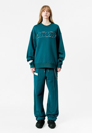 Sudadera de gran tamaño en color azul verdoso con un diseño gráfico, combinada con pantalones holgados a juego que presentan detalles laterales, llevados con calzado negro texturizado.