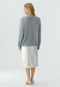 Vrouw die een losse grijze trui, een zilverachtige midi-rok met textuur en beige slingback schoenen met lage hak draagt, staand met haar rug naar de kijker.