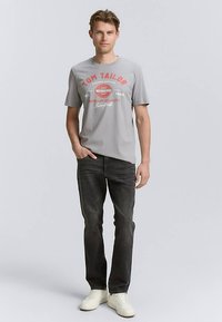 Camiseta de algodón gris con logo impreso en rojo y blanco, combinada con vaqueros oscuros y zapatillas blancas. El ajuste es casual y relajado.