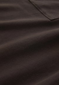 Barna pamutszövet sima textúrával, finom redőzéssel és egy egyszerű négyzet alakú zsebbel a bal felső sarokban.