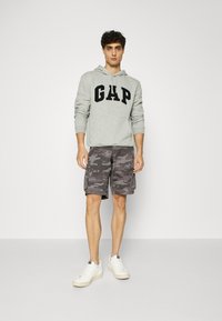 Gray hoodie with "GAP" logo, camo cargo shorts in shades of gray, and white sneakers with textured soles. Model stands against a plain background.