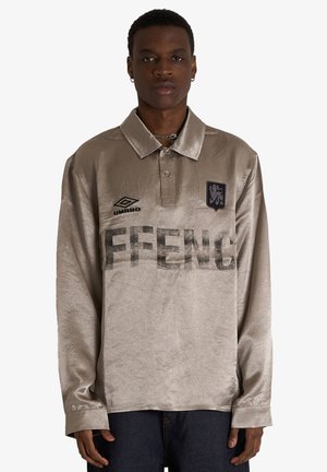 Persona che indossa una polo beige lucida a maniche lunghe con la scritta “FFENCE”, logo Umbro e una toppa nera con emblema di leone.