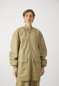 Khaki bomber bunda vyrobená z hladkej syntetickej kože, s celým zipsom, rebrovanými manžetami, dvoma veľkými prednými vreckami a ďalším vreckom na zips.
