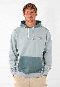 Haut à capuche bleu-gris clair avec une poche vert foncé. Il dispose d'une capuche avec cordon de serrage et d'un petit logo brodé sur la poitrine. Texture de matériau douce.