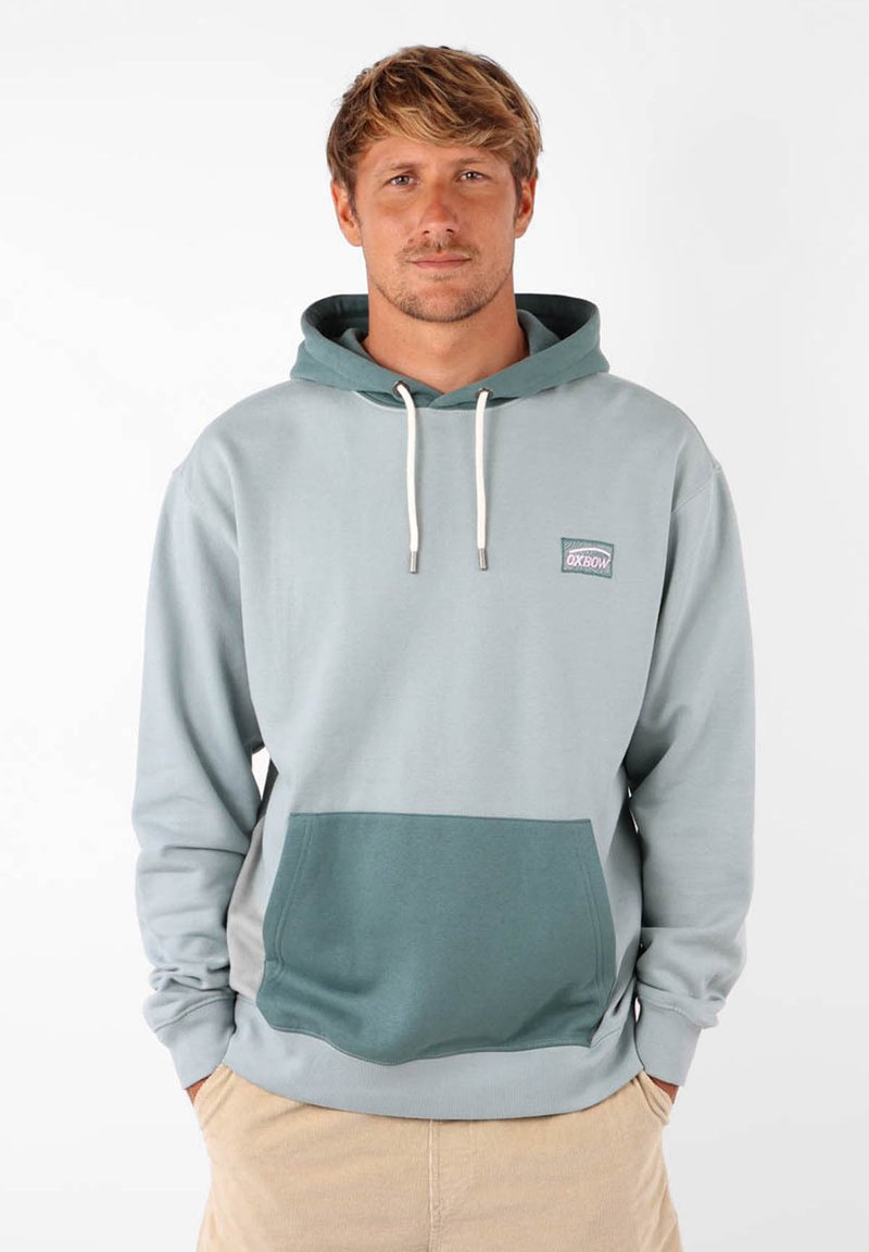 Haut à capuche bleu-gris clair avec une poche vert foncé. Il dispose d'une capuche avec cordon de serrage et d'un petit logo brodé sur la poitrine. Texture de matériau douce.