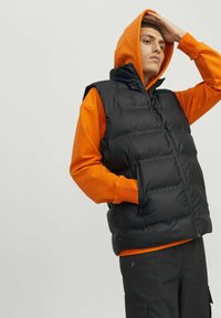 Sort polstret vest over en orange hættejakke, med høj krave, lynlåslommer og en afslappet pasform. Stoffet ser glat og blankt ud.