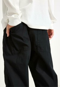 Pantaloni di cotone neri con grandi tasche posteriori, una vestibilità comoda e dettagli di cucitura intricati, abbinati a una camicia bianca a maniche lunghe con texture.