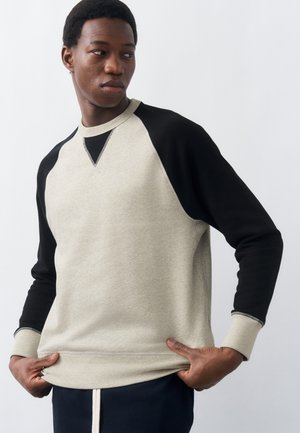 Mees, kes kannab beeži ja musta pika varrukaga sviitrit, hoiab äärelt kinni ja vaatab küljele, taust on hele ja ühtlane.