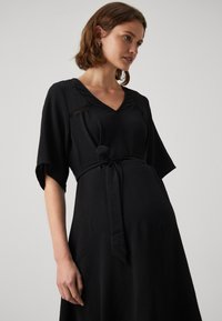 Rochie neagră cu decolteu în V, mâneci largi și curea legată în talie. Prezintă un material texturat și cusături decorative pe umeri.