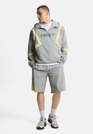 Młody mężczyzna w szaro-żółtej kurtce wiatrówce i pasujących szortach, stoi z rękami w kieszeniach, patrząc na bok.