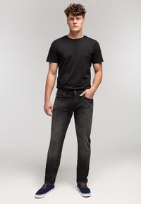 Młody mężczyzna stojący z jedną ręką w kieszeni, ubrany w czarną koszulkę, ciemne dżinsy i granatowe trampki na jednolitym szarym tle.