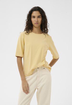 Femeie cu păr creț și întunecat, purtând un top larg cu mâneci scurte de culoare galben pal și pantaloni bej deschis, stând cu o mână în buzunar.